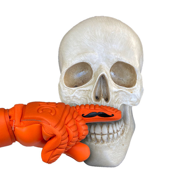 White skull with a bright orange soccer goalie glove that has a mustache printed on the inside index finger. The mustache from the glove is being placed over the skull where the mustache would go, above the upper lip of the skull. The color of the soccer goalie glove is for Halloween. 